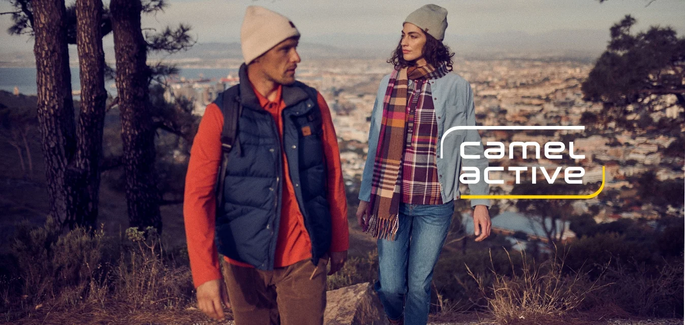 Due persone stanno camminando su un sentiero di montagna, indossando una giacca blu, una sciarpa a quadri, un berretto beige e un gilet scuro sopra una maglietta arancione.