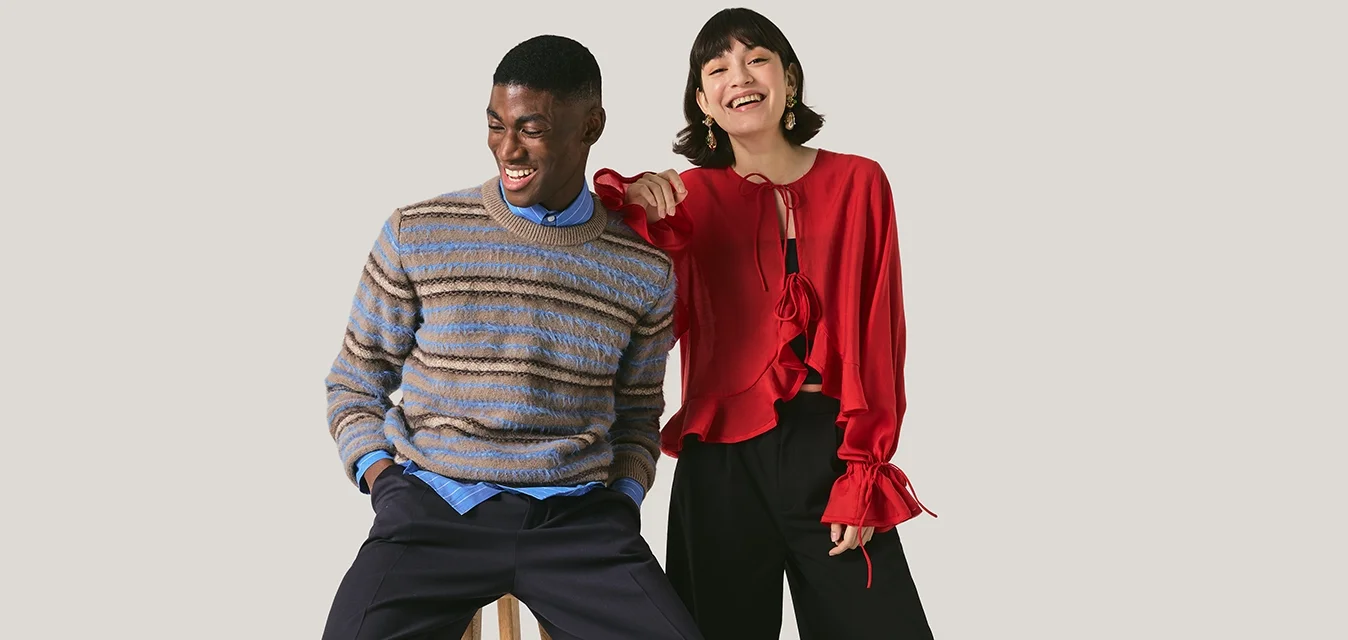 Uomo con maglione a righe seduto su uno sgabello, donna con camicetta rossa in piedi accanto a lui, sorridente e che indica in avanti.