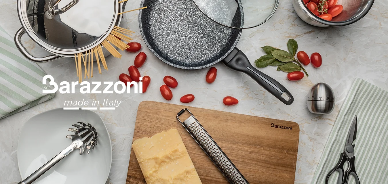 Pentola in acciaio inossidabile con spaghetti, padella antiaderente con coperchio di vetro, grattugia argentata, tagliere di legno, pomodorini, erbe aromatiche e utensili.