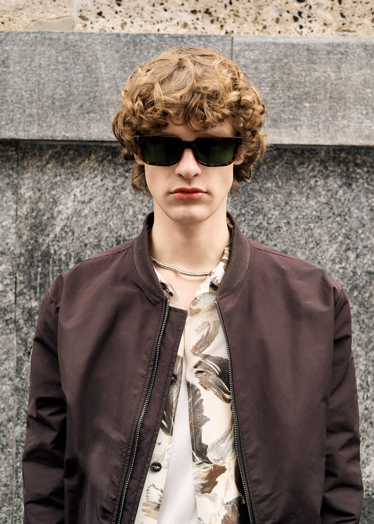 Young man with curly hair wearing sunglasses and a brown bomber jacket over a patterned shirt against a stone wall