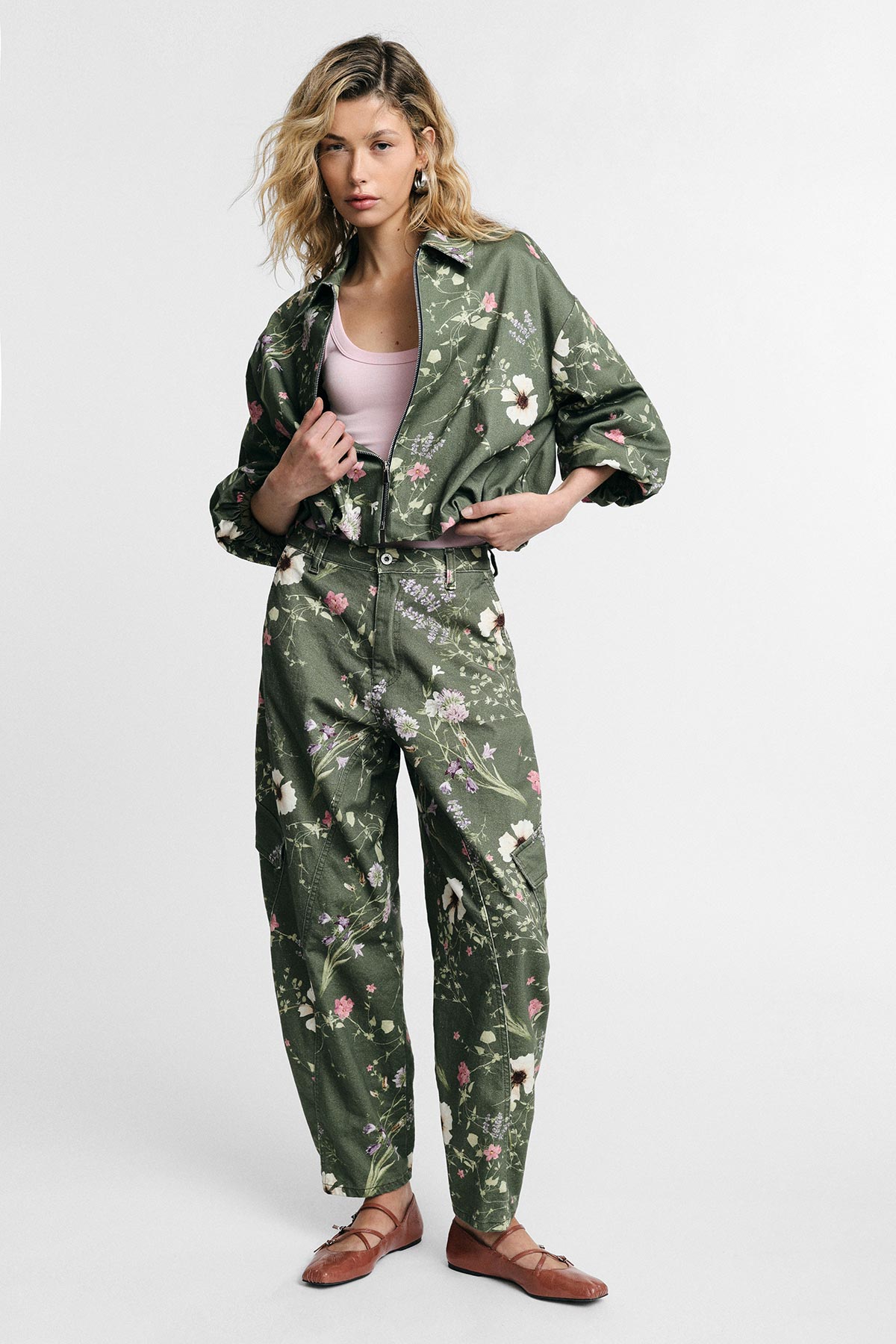 Young woman modelling green floral co-ord set with matching jacket and trousers, pink camisole and brown ballet flats on white background