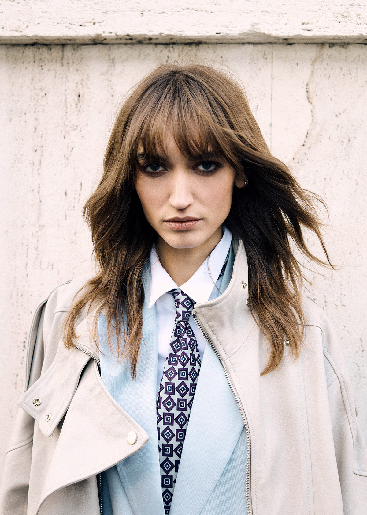 Female model with brown hair and bangs wearing beige coat, light-blue blazer and patterned tie against concrete wall