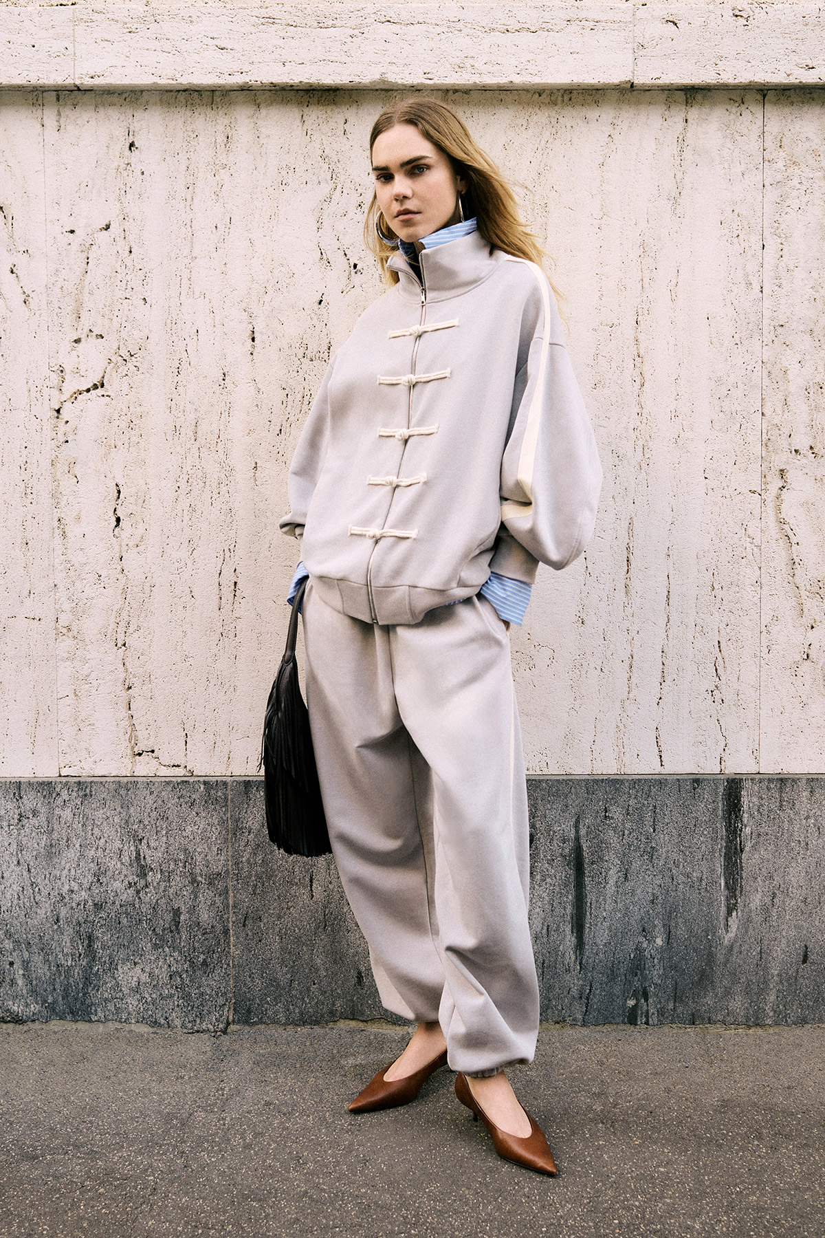 Woman standing against a stone wall wearing oversized beige toggle-jacket and trousers, blue cuffs, brown pointed flats and a black fringe bag
