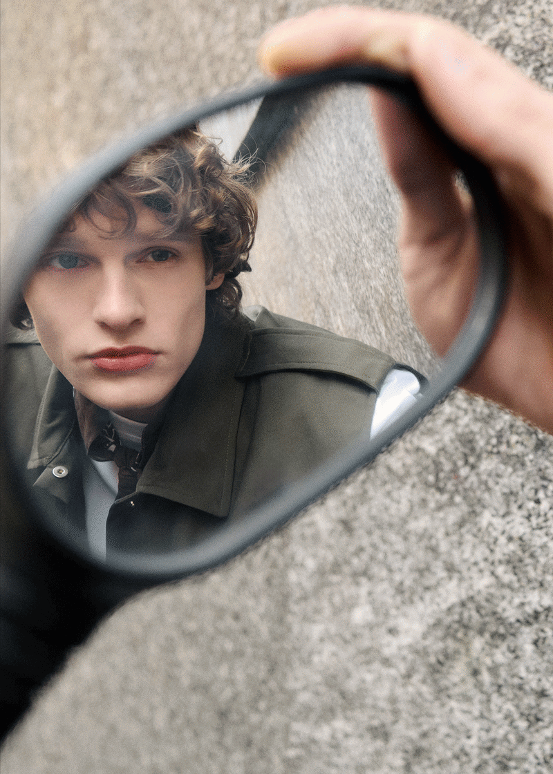 Triptych fashion collage: close-up face in a handheld mirror; monochrome full-length figure by a concrete wall; model in tan jacket and cream knit