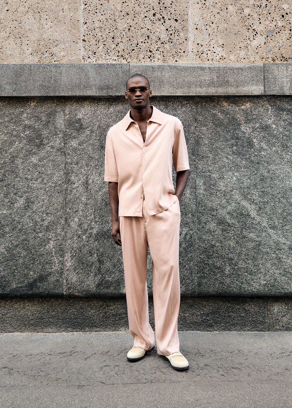 Man in blush-pink relaxed shirt and matching wide trousers with beige shoes, standing against a textured stone wall