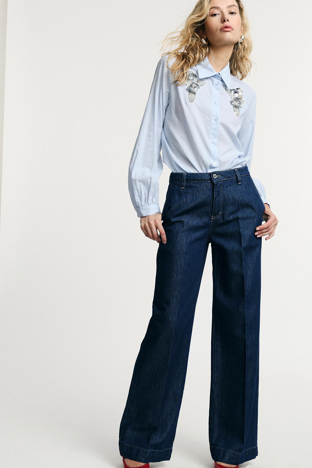Woman in light-blue embroidered blouse and wide-leg dark denim jeans, hands in pockets against plain studio background