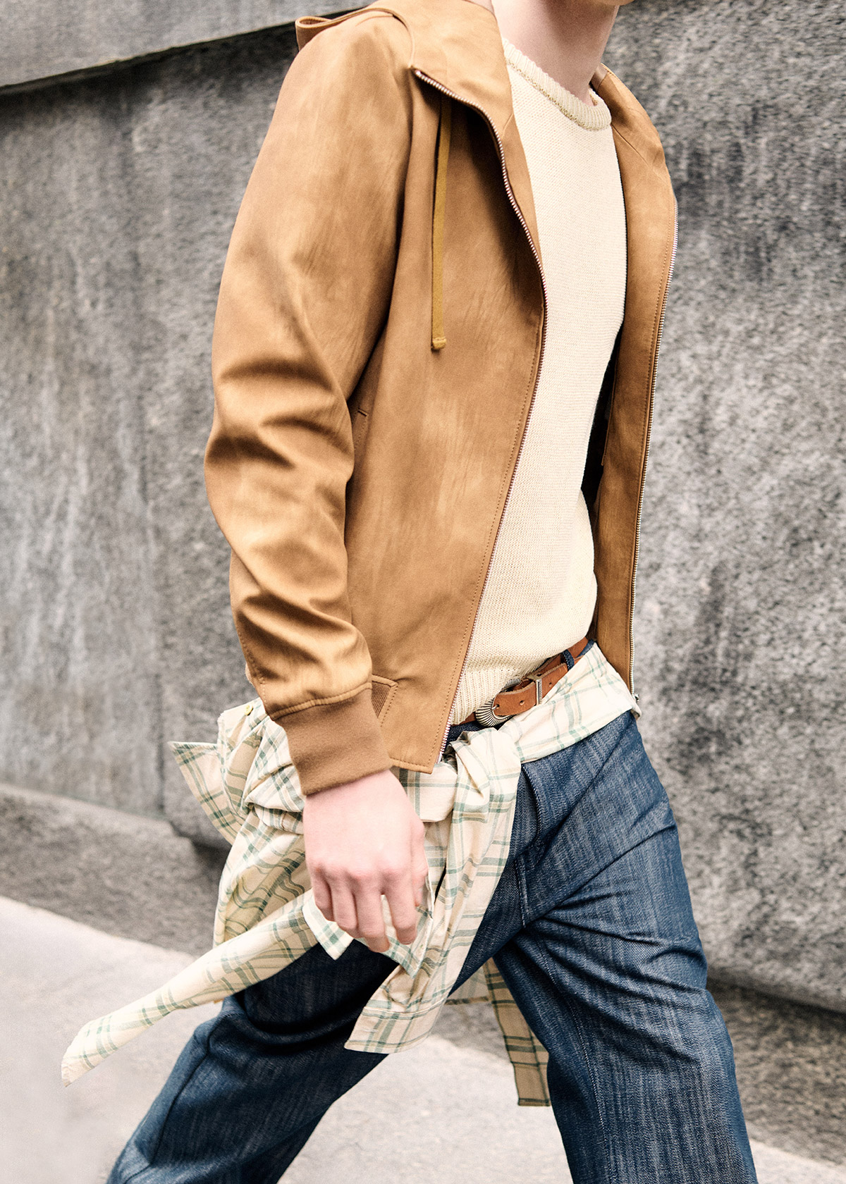 Man walking by concrete wall wearing tan suede jacket, cream knit sweater, blue jeans and a plaid shirt tied around the waist