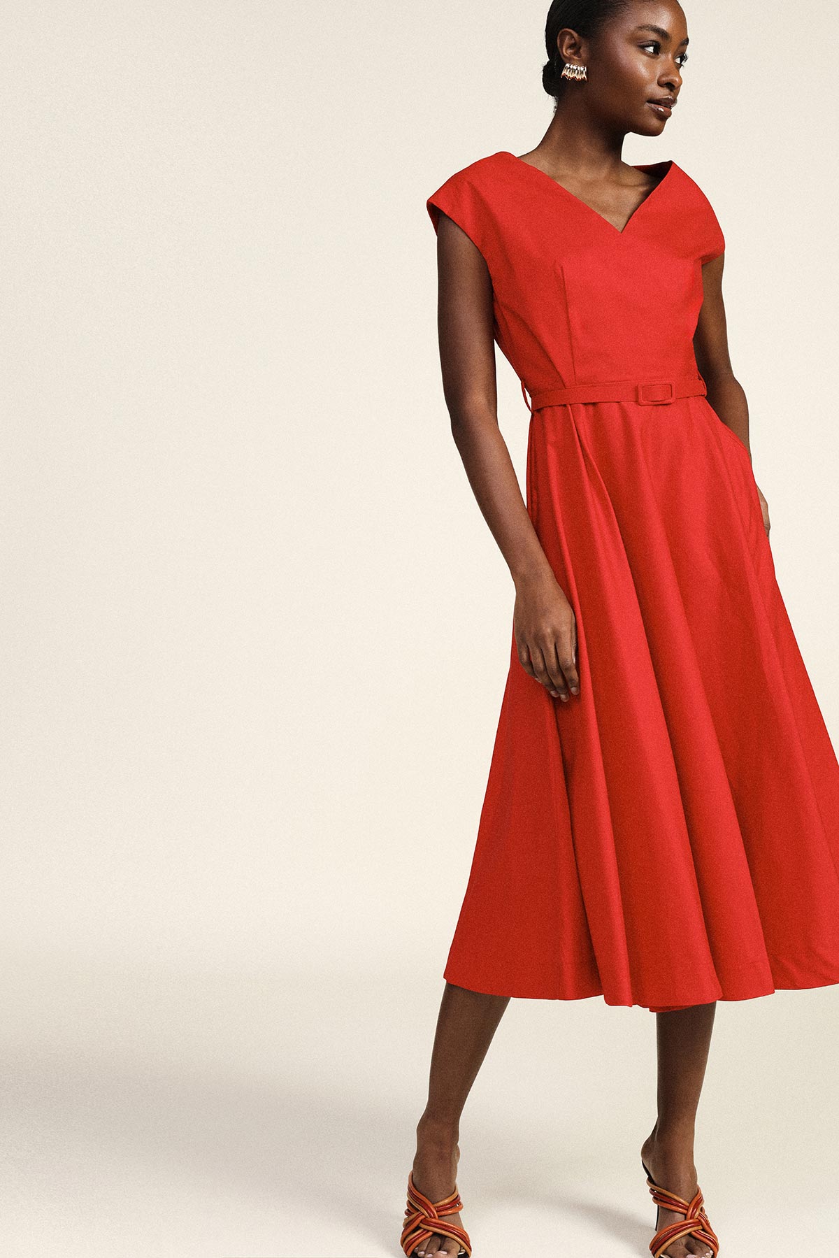 Woman modelling red V-neck belted midi dress with cap sleeves and strappy sandals on neutral studio background