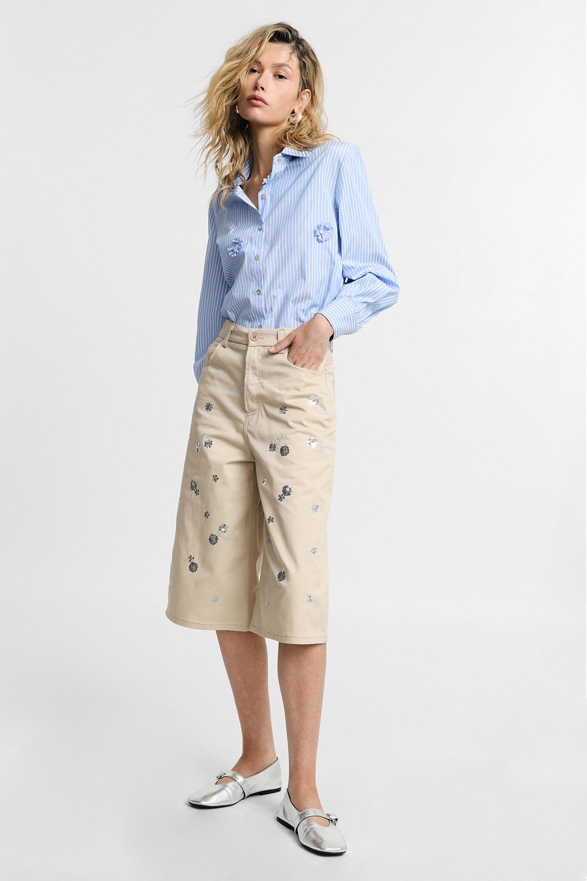 Female model wearing a blue striped shirt, beige embellished culotte shorts and silver loafers, standing against a white background