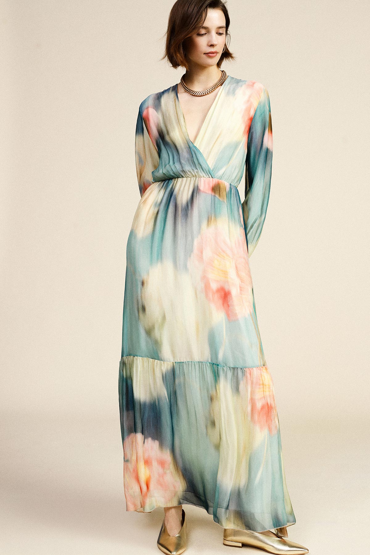 Model wearing pastel watercolour floral V‑neck long-sleeve maxi dress with gold necklace and metallic shoes on neutral background