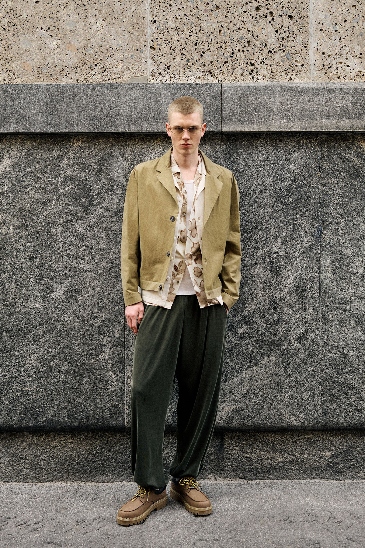 Man in beige jacket, patterned shirt, loose olive trousers and brown shoes against textured stone wall, men's casual fashion