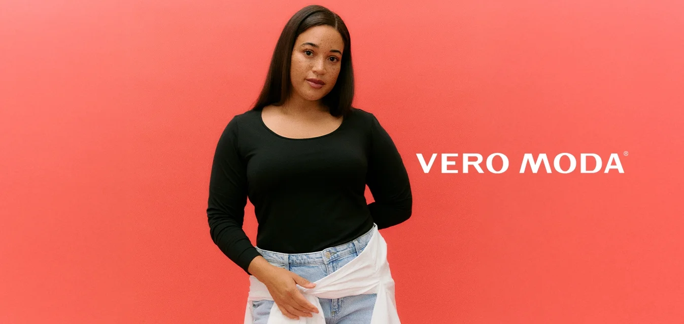 Donna con lunghi capelli scuri che indossa una maglia nera a maniche lunghe, jeans azzurri chiari e una camicia bianca annodata in vita, su uno sfondo corallo.