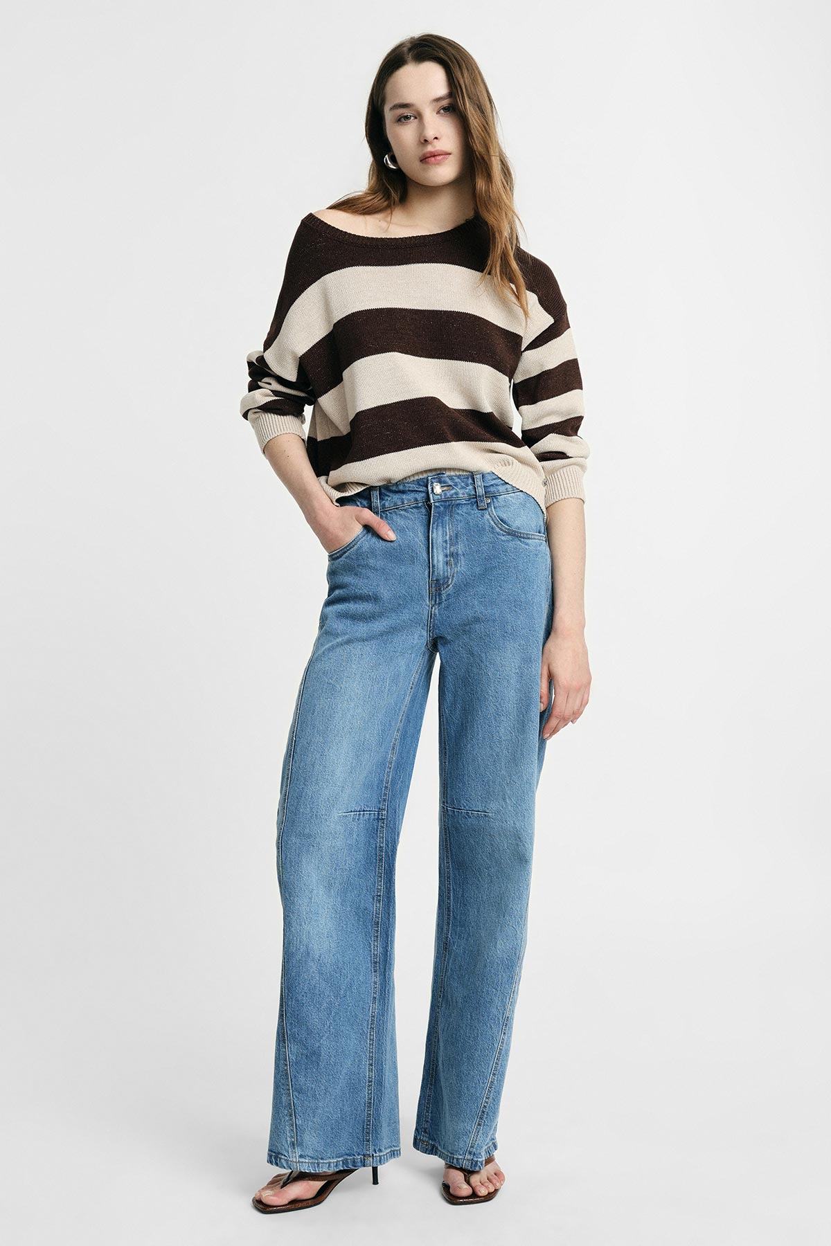 Young woman model wearing cream and dark brown striped jumper, high-waisted wide-leg blue jeans and sandals, studio background