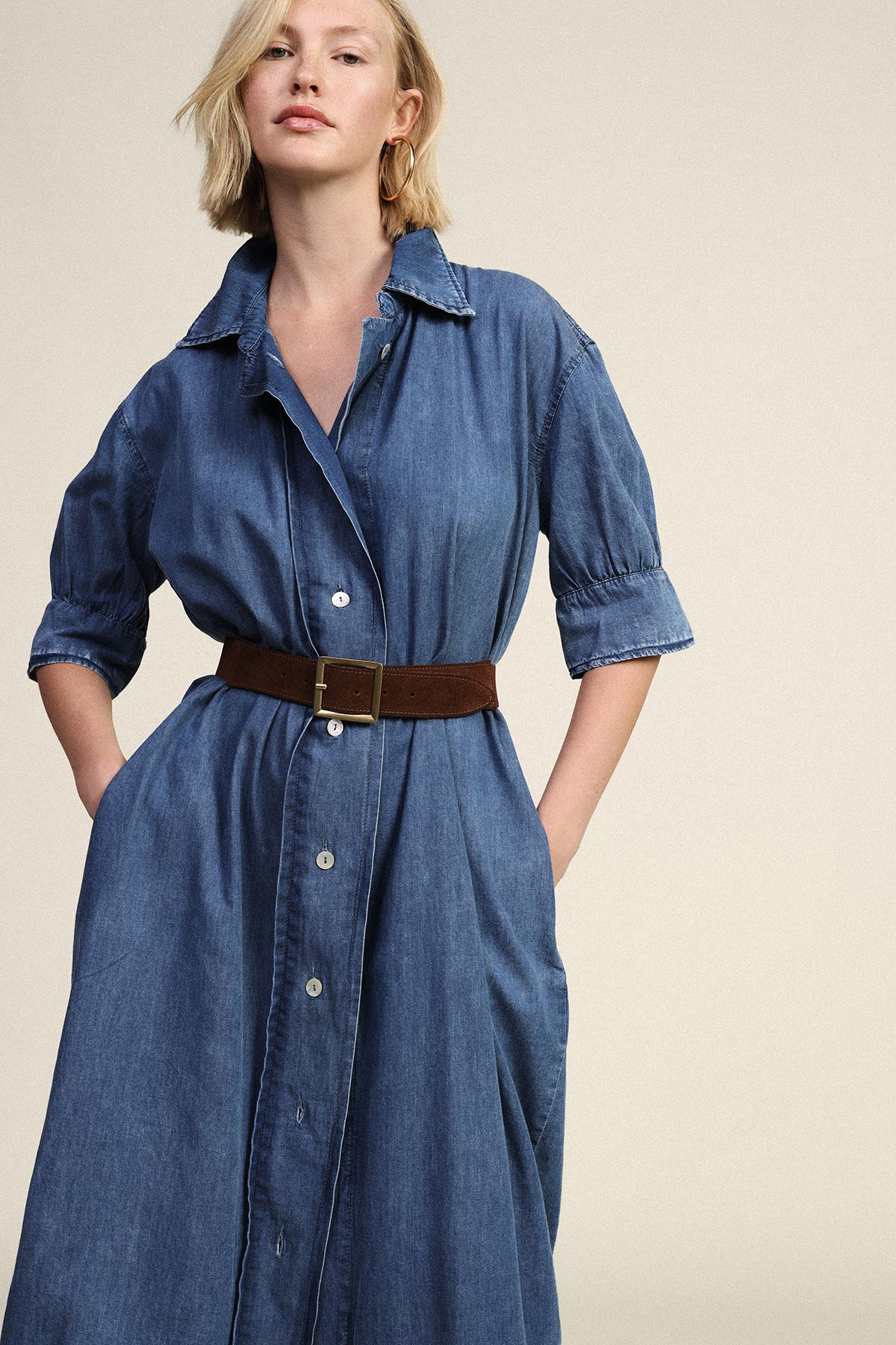Model wearing a knee-length blue denim shirt dress with rolled sleeves and a brown suede belt, hands in pockets, neutral background