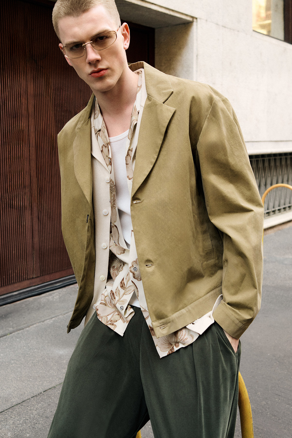 Young man wearing khaki blazer, floral shirt and sunglasses with hands in pockets standing on city pavement