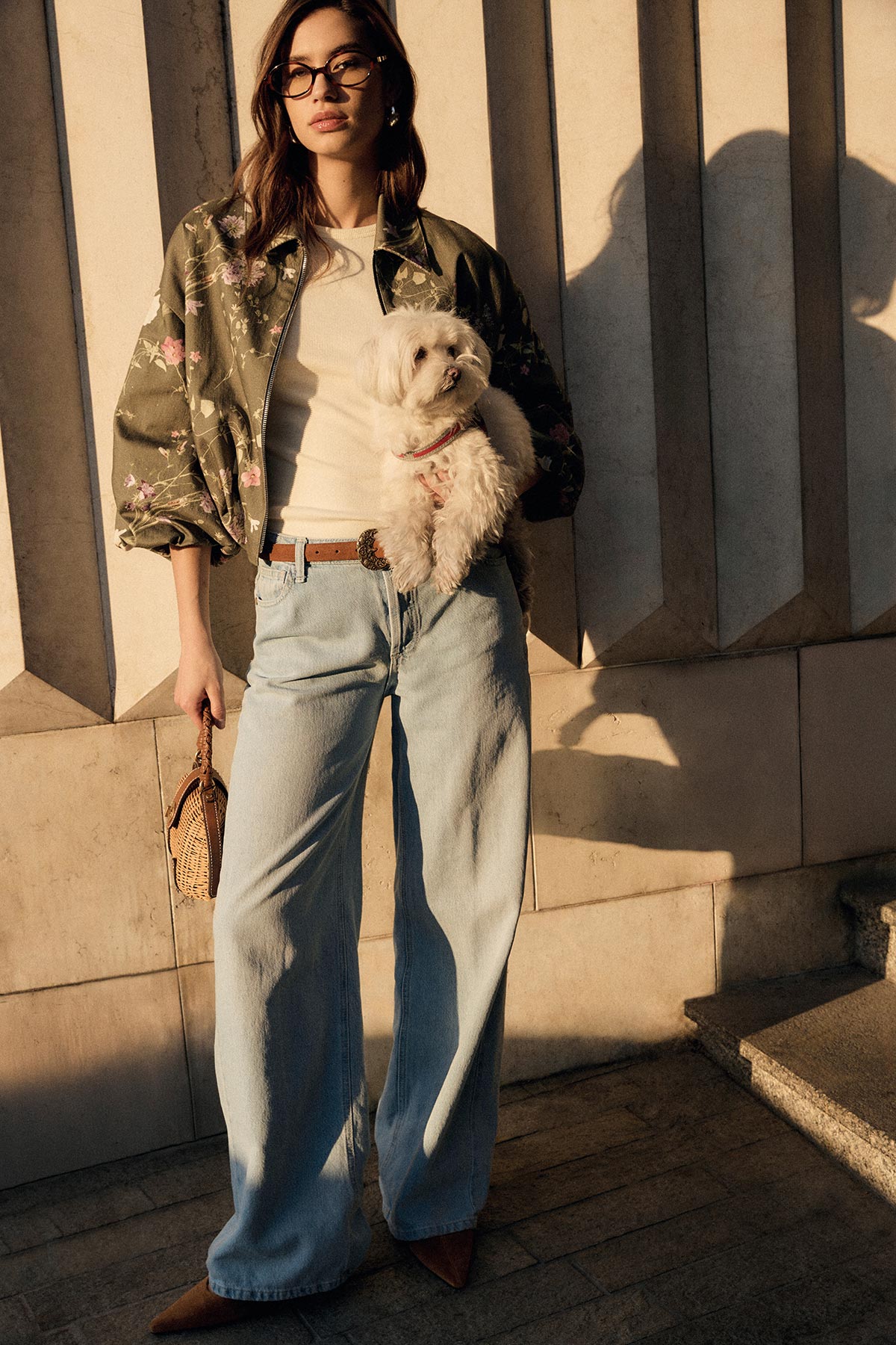 Woman in floral bomber jacket, white top and wide‑leg jeans holding a small white dog with a wicker bag by a sunlit stone wall