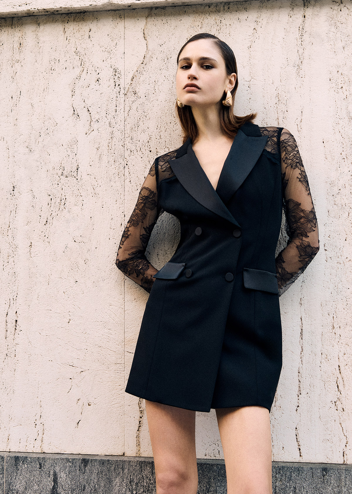 Fashion model in black double-breasted blazer dress with lace sleeves and gold earrings, leaning against a textured wall