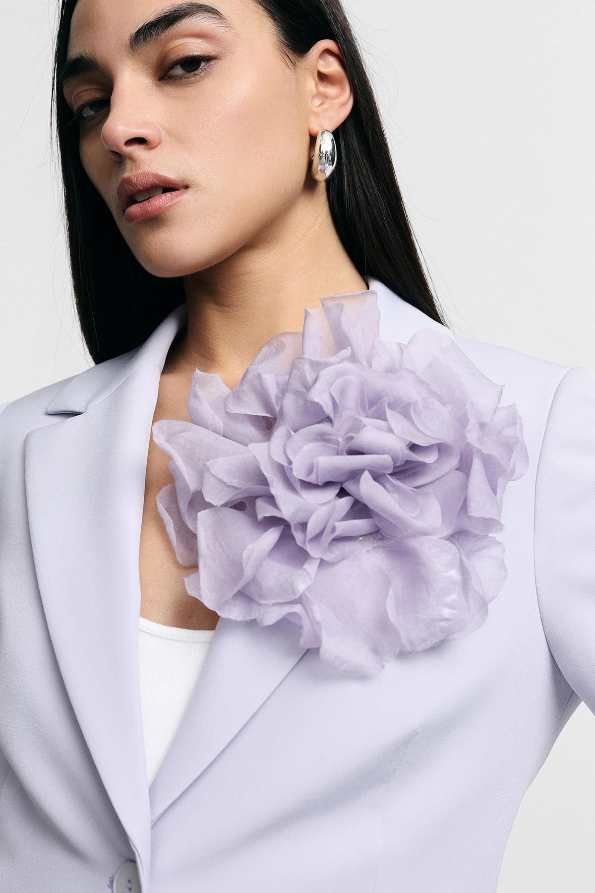 Close-up of a woman in a lilac blazer wearing an oversized fabric flower corsage and silver hoop earring, fashion portrait