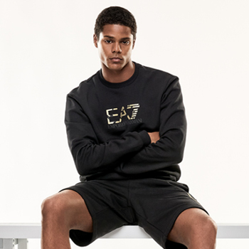 Male model seated on bench wearing black EA7 Emporio Armani sweatshirt and black shorts, arms crossed