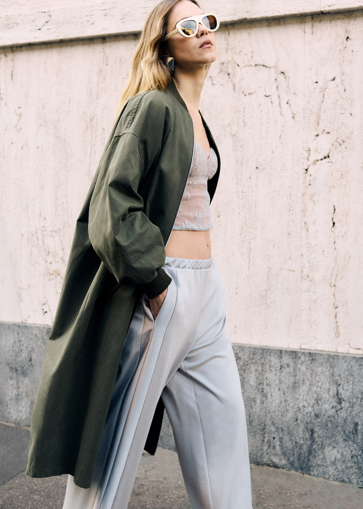 Woman in olive oversized coat, lace crop top and light tailored trousers wearing white sunglasses, walking past a pale textured wall