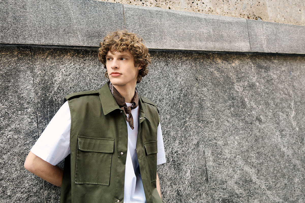 Young man with curly hair in olive utility vest, white T‑shirt and brown neckerchief leaning on grey stone wall, street style