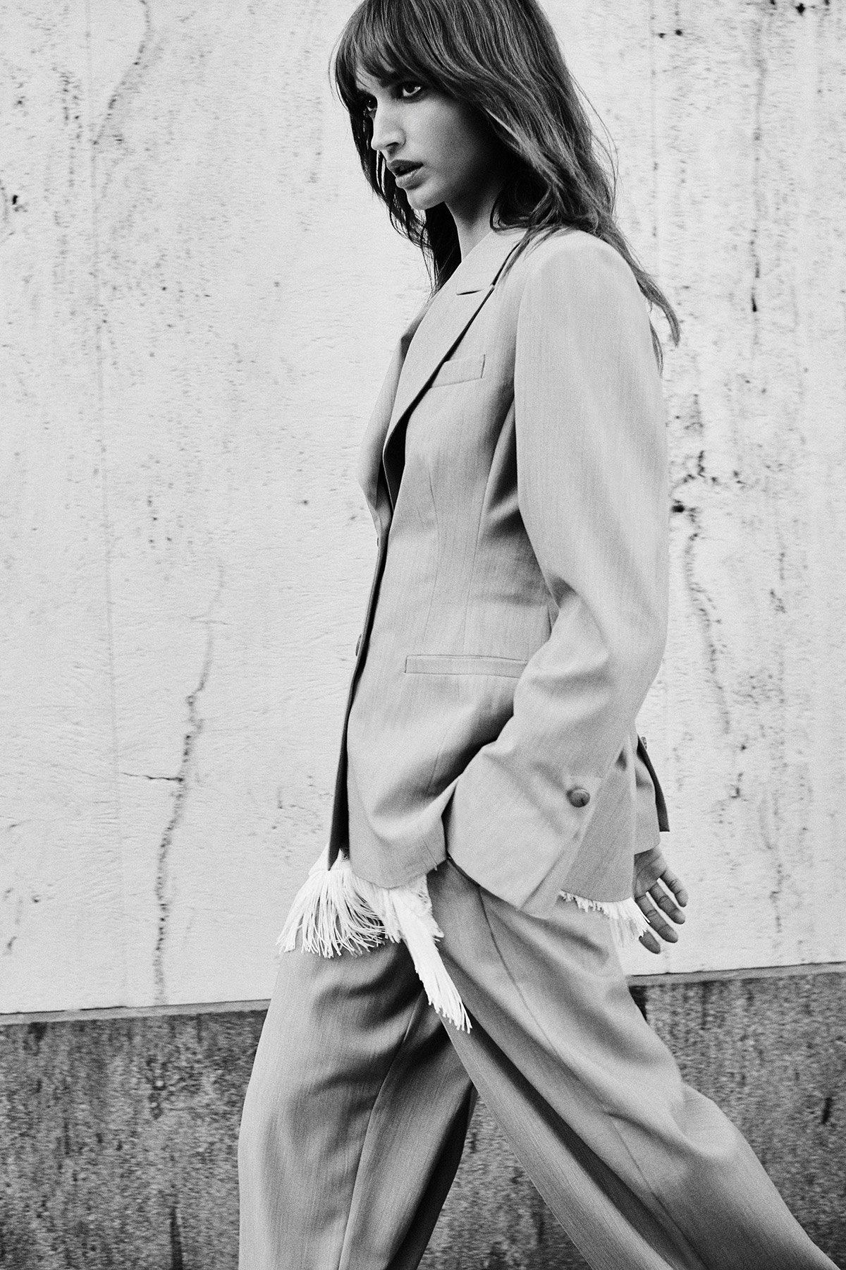 Black-and-white photo of a woman in a tailored suit walking past a textured concrete wall