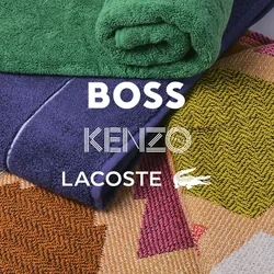 Mucchio di asciugamani piegati, uno verde arrotolato, uno viola piegato, posati su un tappeto colorato con motivo geometrico e bordi frangiati.