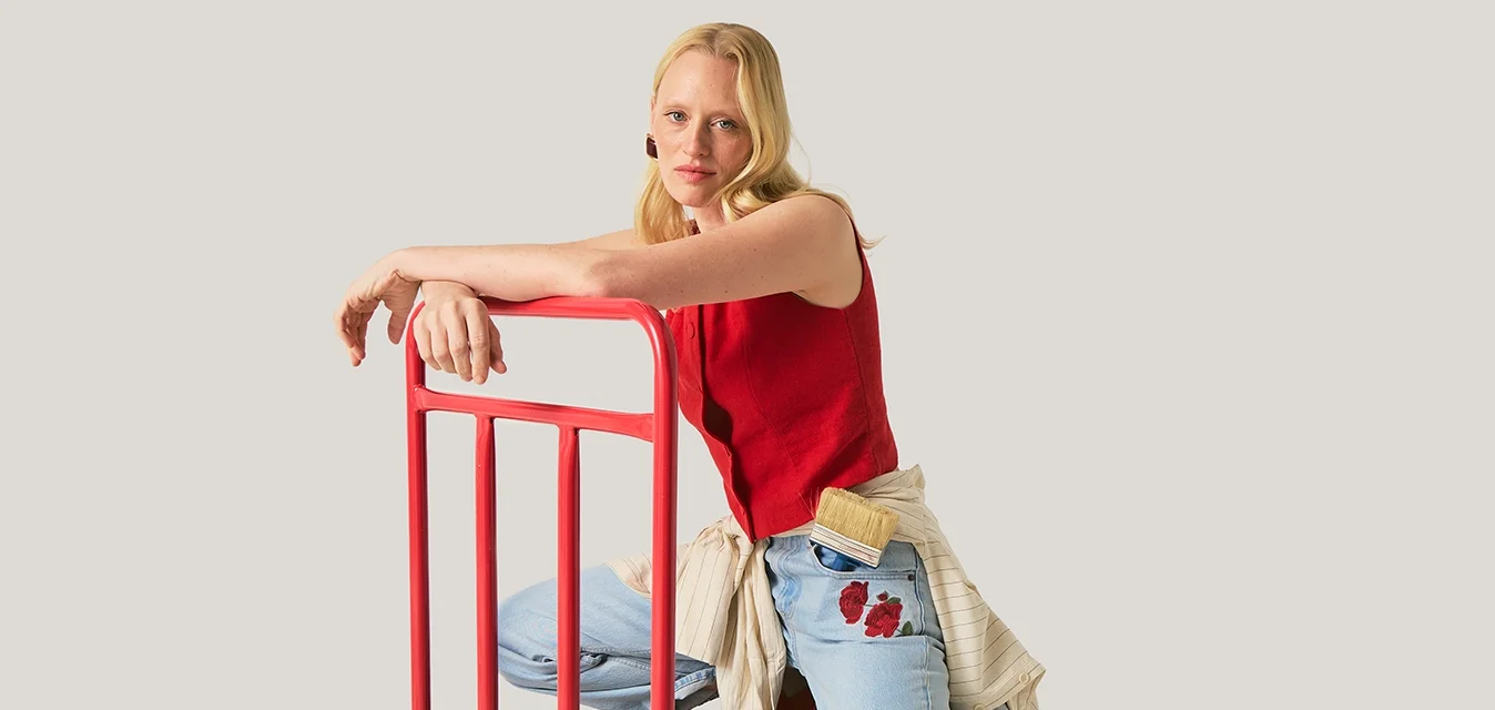 Donna con top rosso e jeans inginocchiata su un carrello piattaforma nero con manico rosso su sfondo neutro.