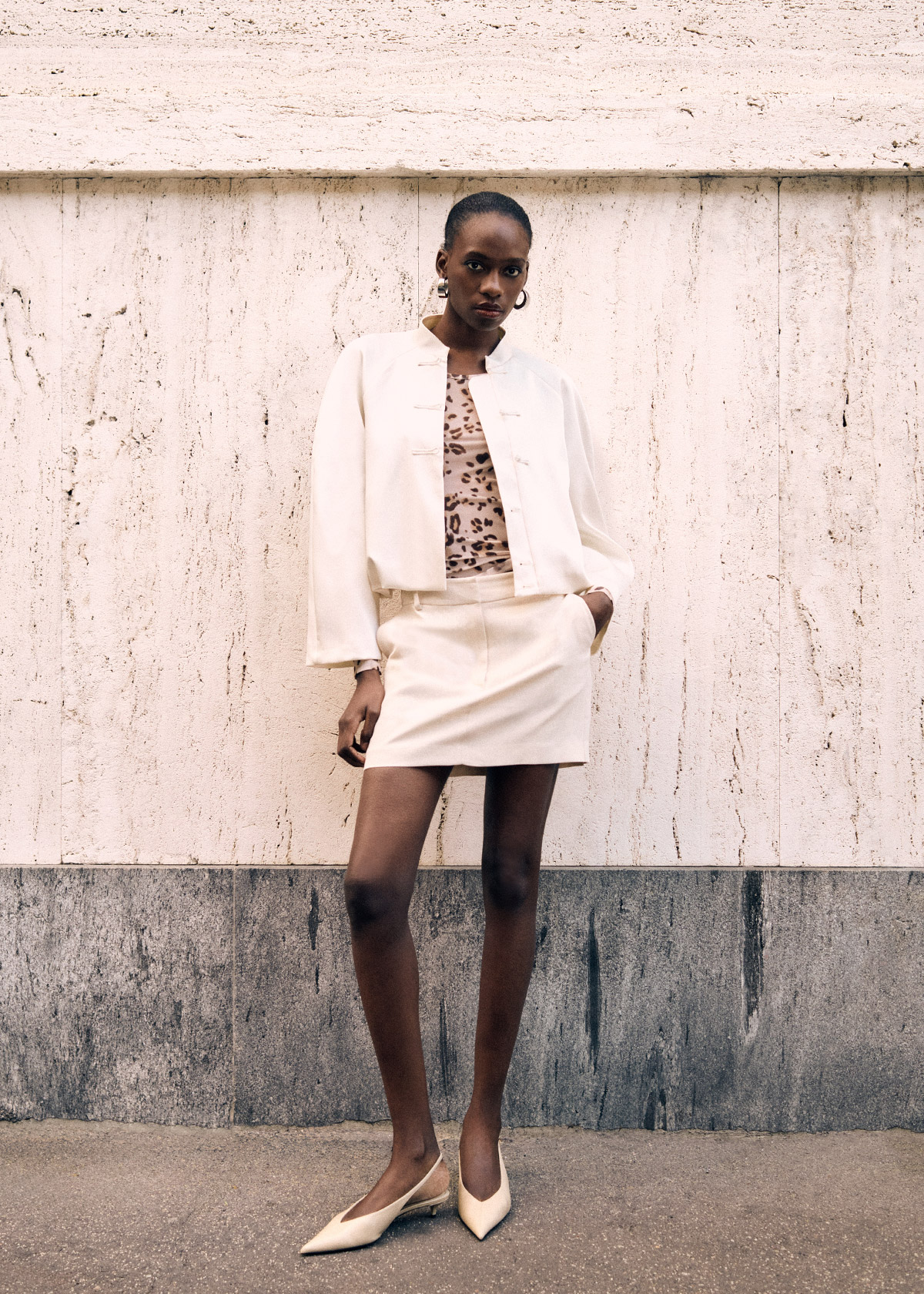 Female model in cream blazer and matching mini skirt with leopard-print top and pointed heels posing against a textured stone wall