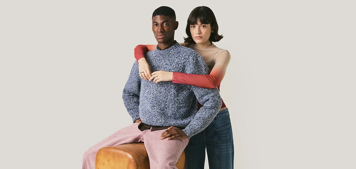 Un uomo con un maglione blu e pantaloni rosa è seduto su una panca da palestra, mentre una donna con una maglietta rossa e jeans a gamba larga si trova dietro di lui, abbracciandolo.