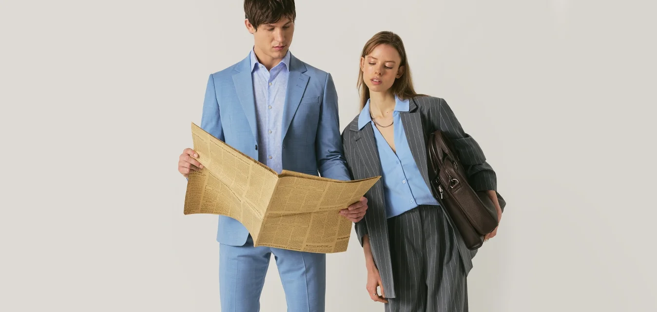 Uomo in abito azzurro chiaro che tiene un giornale, donna in abito grigio a righe con camicia azzurra che si china verso il giornale, tenendo una borsa marrone.