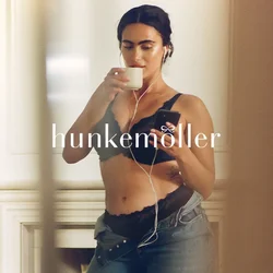 Bralette in pizzo nero con bordi smerlati, jeans in denim a vita alta e auricolari. Tiene una tazza bianca con una mano e uno smartphone con l'altra.