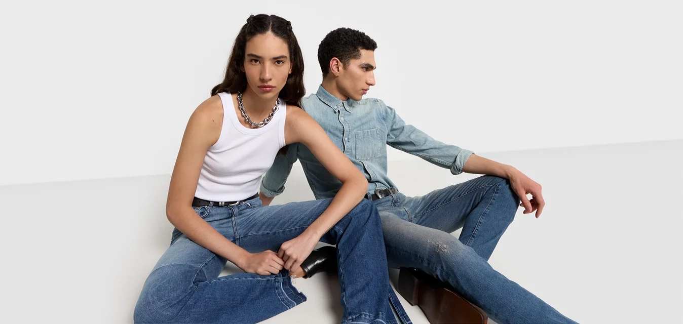 Due persone sedute su una superficie bianca. Una indossa una canotta bianca e jeans blu, l'altra una camicia di denim chiaro e jeans scuri.