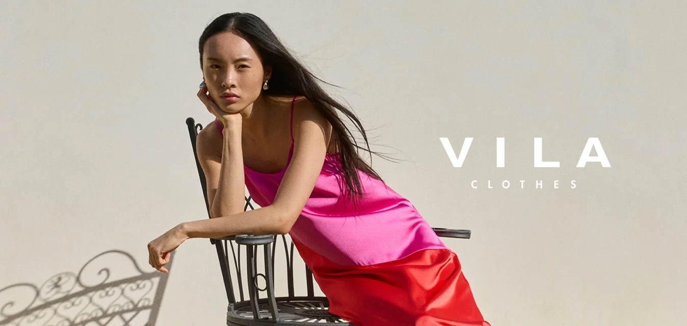 Donna con abito rosa e rosso appoggiata a una sedia di metallo nero su pavimento piastrellato contro una parete chiara e liscia.