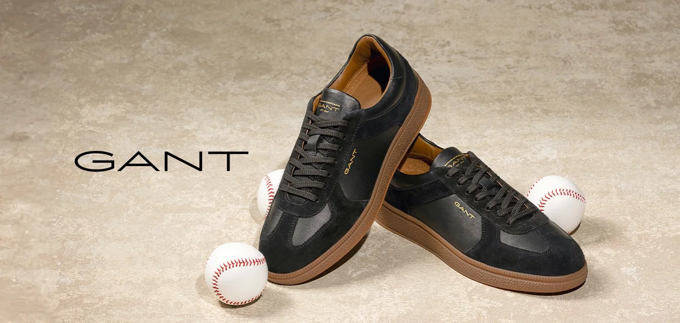 Sneaker nere in pelle con dettagli in suede, suole in gomma e rifiniture marroni. Posizionate vicino a due palline da baseball su uno sfondo testurizzato.