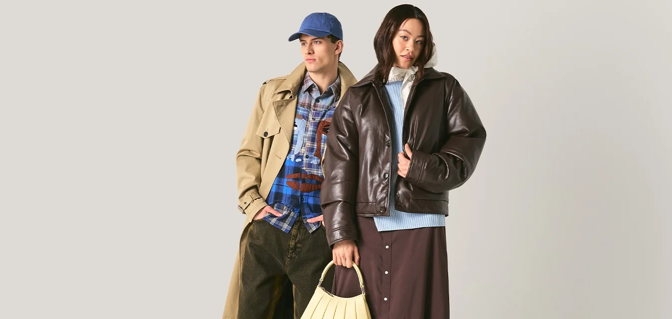 Due persone sono in piedi davanti a uno sfondo semplice; una indossa un trench e un cappello, l'altra una giacca di pelle e una gonna, tenendo una borsa beige.