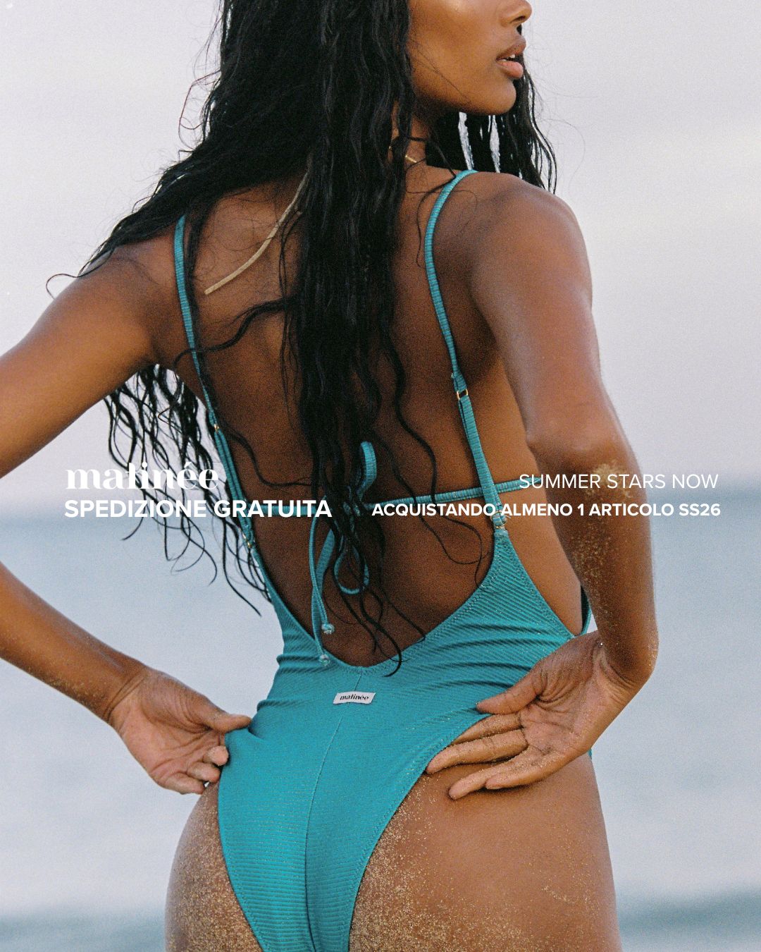 Back view of a woman in a turquoise one-piece swimsuit standing on a sandy beach with long wet hair, summer fashion shot