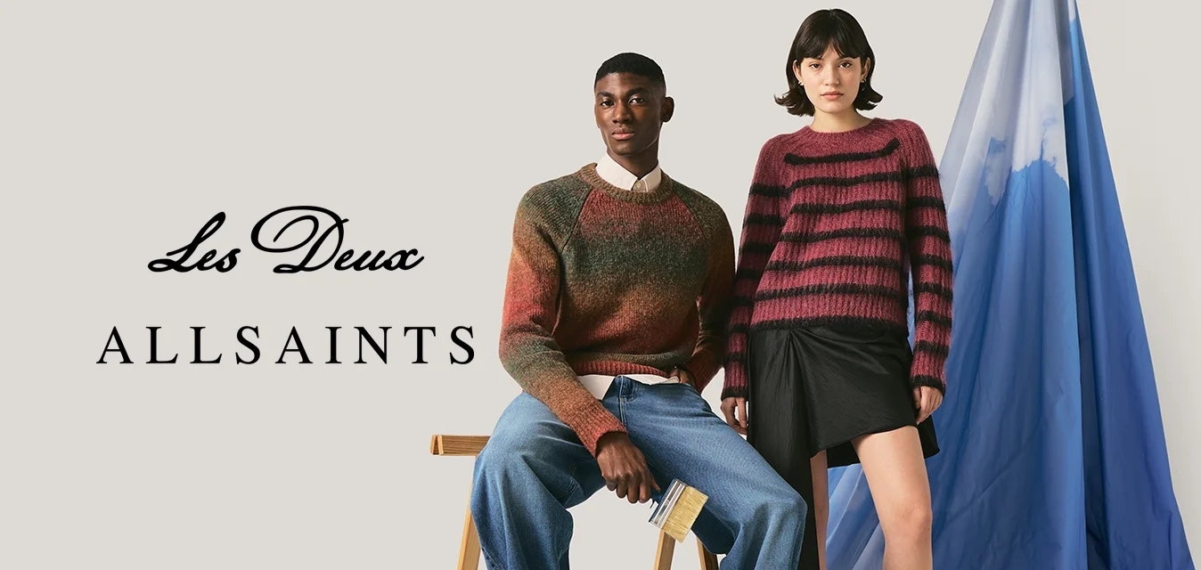 Due modelle, una seduta su uno sgabello indossa un maglione marrone e jeans, l'altra in piedi con un maglione a righe e una gonna nera accanto a un tessuto blu drappeggiato.