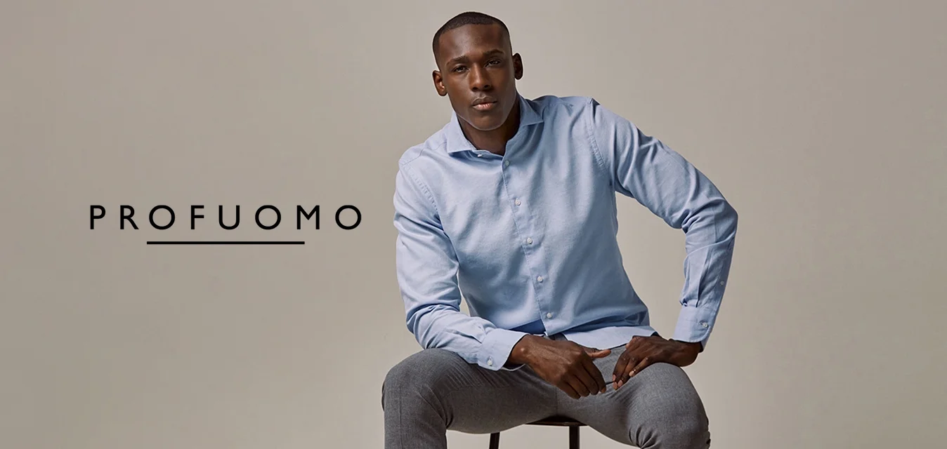 Camicia azzurra chiara con bottoni, pantaloni grigi e sneakers grigie. Il modello è seduto su uno sgabello nero con le mani sulle ginocchia, contro uno sfondo neutro.