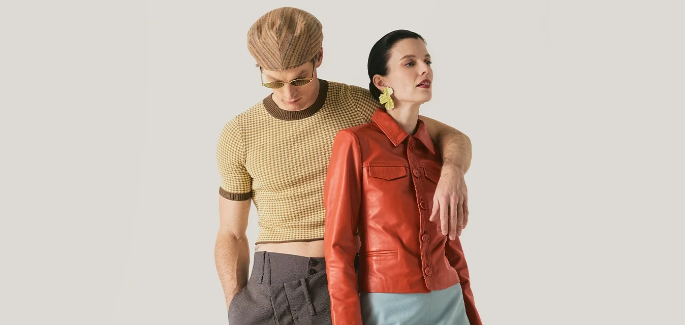 Uomo con cappello marrone pied de poule e camicia corta con pantaloni grigi, braccio intorno a donna con giacca di pelle rossa, gonna blu e stivali beige.