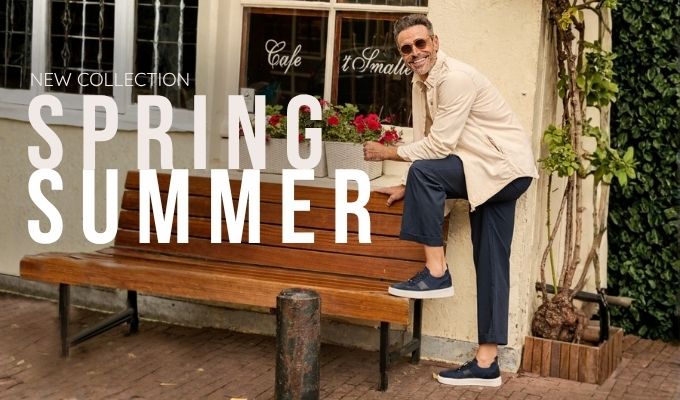 Man in beige jacket, navy pants and sneakers leaning on bench by cafe window with flower box — spring summer fashion
