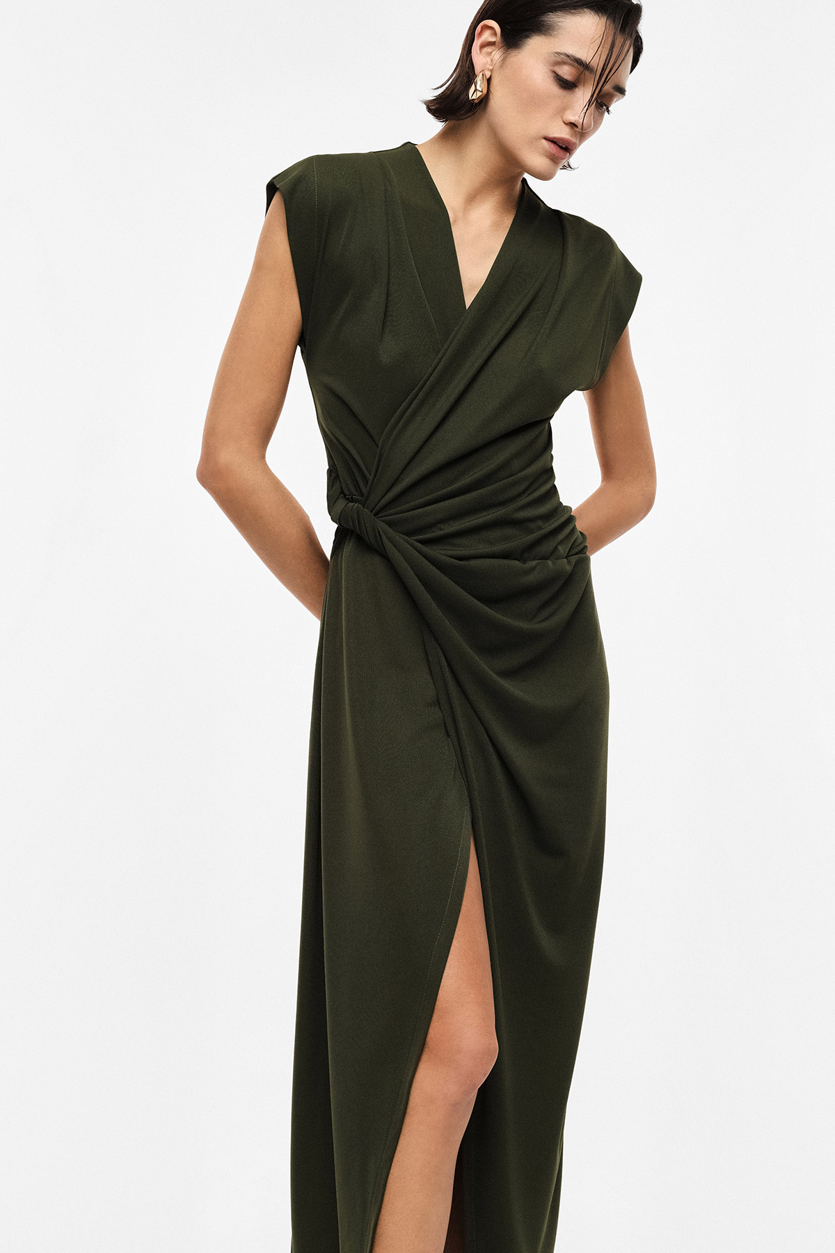Woman modelling olive-green wrap midi dress with knot waist and thigh slit, cap sleeves and gold hoop earrings on white background