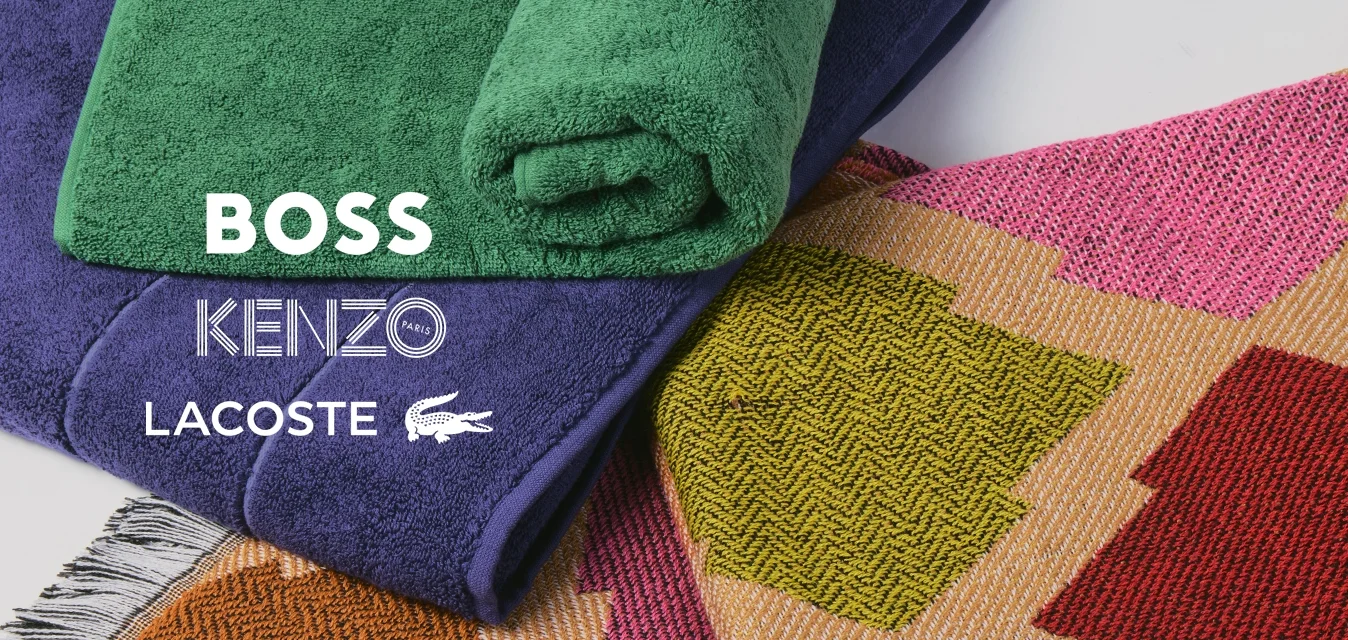 Mucchio di asciugamani piegati, uno verde arrotolato, uno viola piegato, posati su un tappeto colorato con motivo geometrico e bordi frangiati.