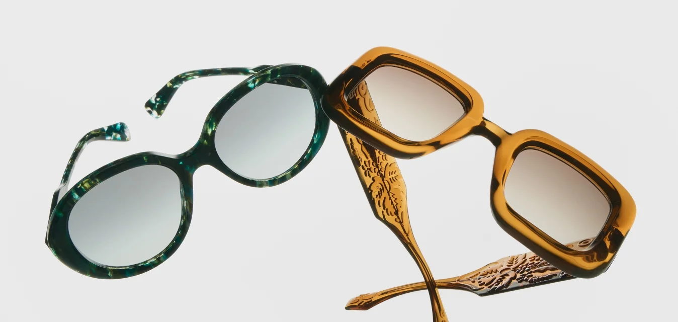 Due occhiali da sole, uno con montatura rotonda verde e l'altro con montatura quadrata arancione, entrambi con aste decorate su uno sfondo bianco.