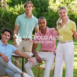 Quattro persone indossano polo pastello e pantaloni chiari, circondate da vegetazione. Una sedia in rattan e un martello di legno chiaro sono nelle vicinanze.