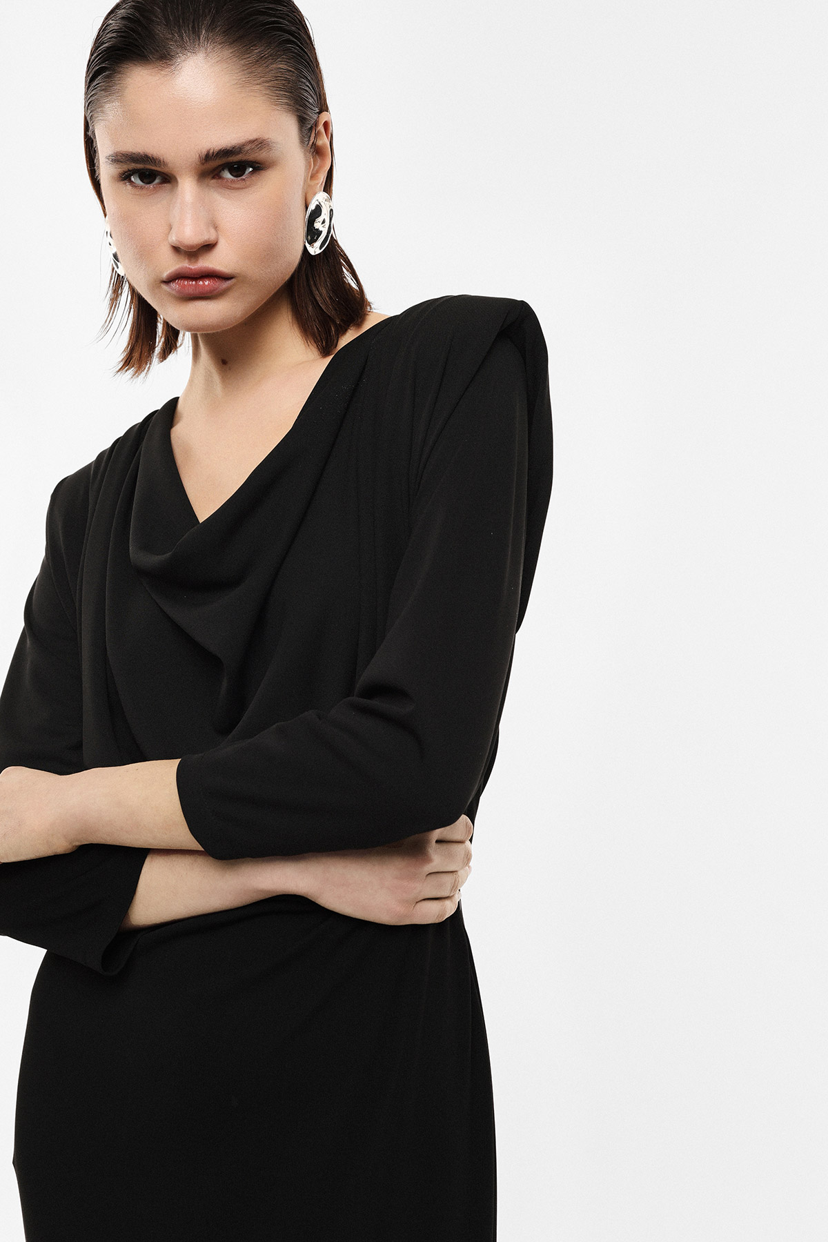 Woman wearing a black draped dress with three-quarter sleeves and silver statement earrings on white background