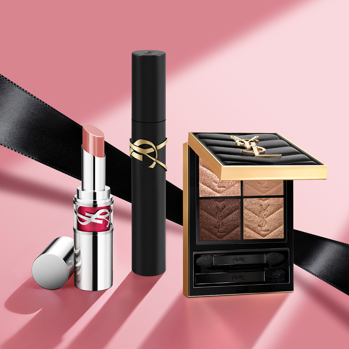 Un rossetto, un mascara e una palette di ombretti YSL sono elegantemente disposti su uno sfondo rosa con un nastro nero.