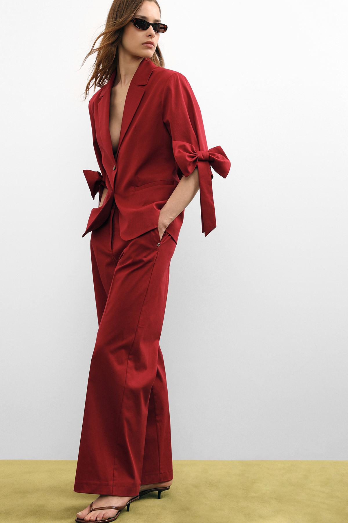 Woman in deep-red tailored blazer with bow-tie sleeves and wide-leg trousers, wearing sunglasses, hands in pockets
