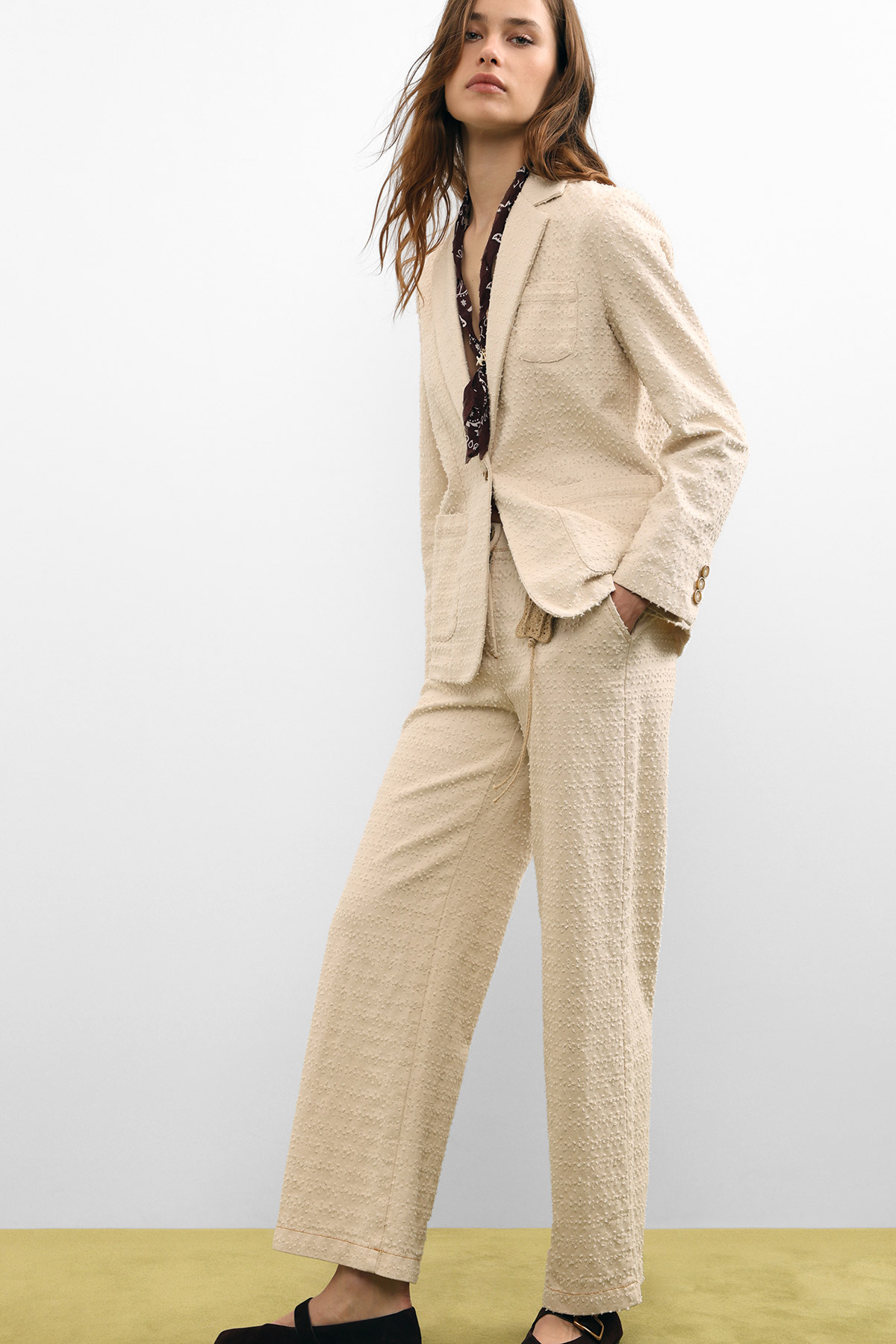 Woman modelling beige textured blazer and wide-leg trousers with dark patterned blouse and black sandals, hands in pocket