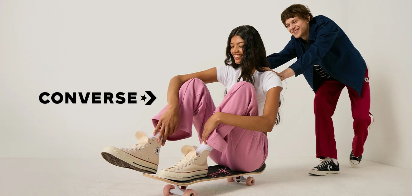 Giovane donna con pantaloni rosa seduta su uno skateboard mentre un uomo sorridente la spinge da dietro in uno spazio studio bianco.